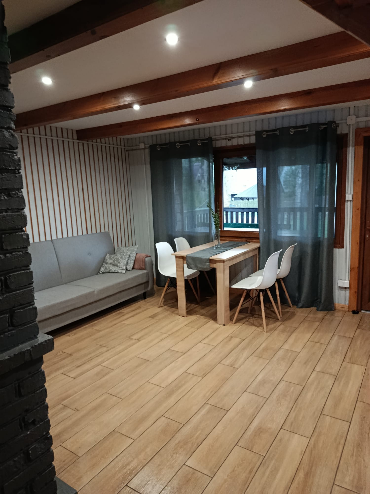 Salon z sofą, stołem jadalnym i dużym oknem z zasłonami, widok na taras.