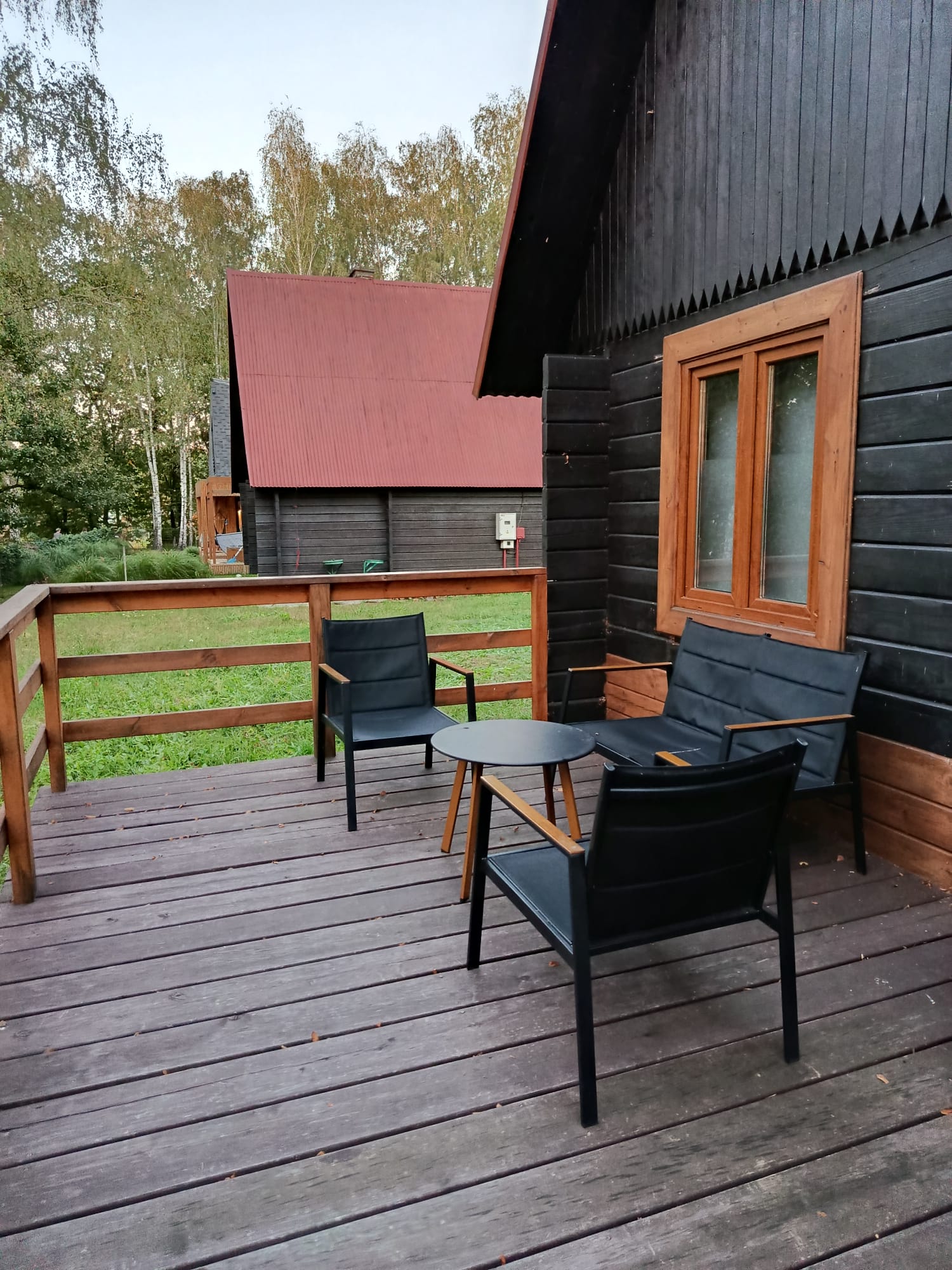Taras z drewnianą podłogą, stolikiem i wygodnymi krzesłami przed domkiem letniskowym.