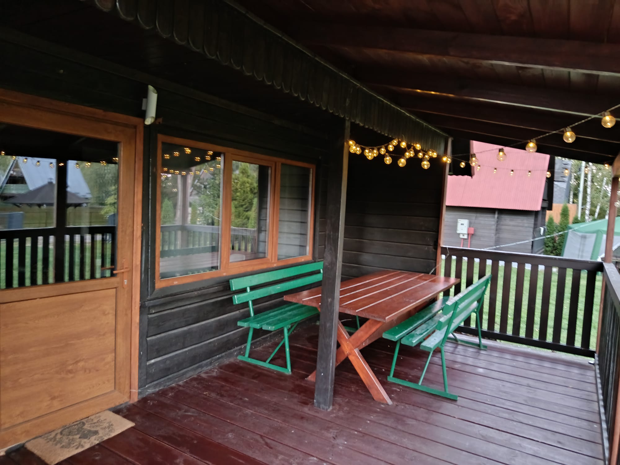 Zadaszony taras z drewnianym stołem i zielonymi ławkami, ozdobiony girlandami świetlnymi.