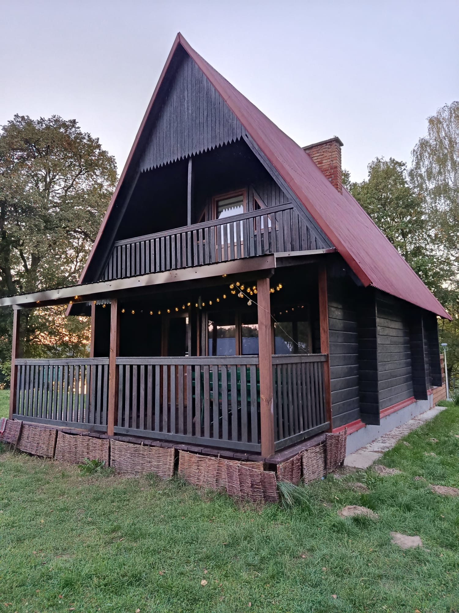 Drewniany domek letniskowy z czerwonym dachem i balkonem na piętrze, w otoczeniu zieleni.