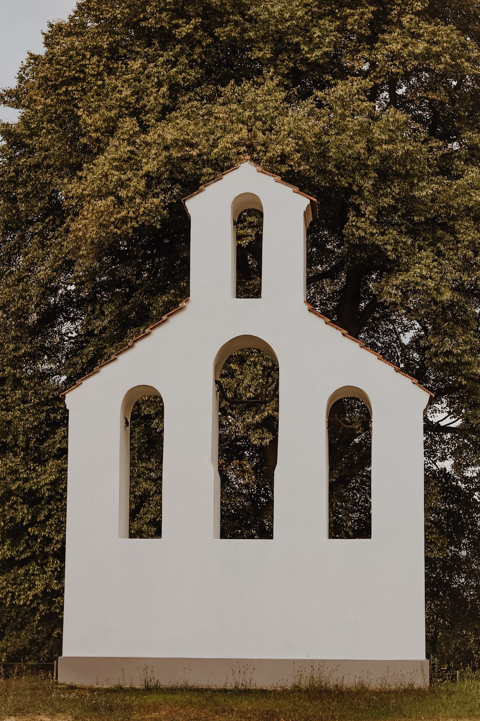 Biała kapliczka polowa stojąca w cieniu dużego drzewa na terenie ośrodka.